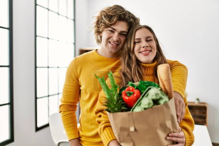 Téléchargez les photos : Jeune couple caucasien tenant épicerie sac en papier s'embrassant à la maison - en image libre de droit