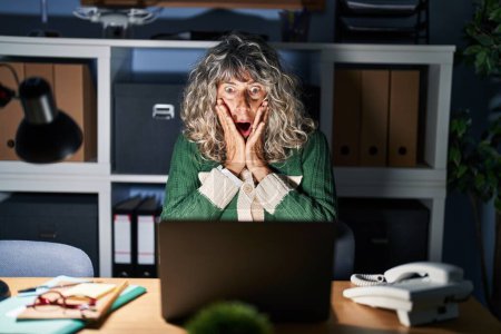 Foto de Middle age woman working at night using computer laptop afraid and shocked, surprise and amazed expression with hands on face - Imagen libre de derechos