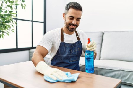 Foto de Hombre hispano joven limpiando mesa usando pulverizador en casa - Imagen libre de derechos