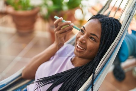 Photo for African american woman listening audio message by smartphone lying on hammock at home terrace - Royalty Free Image