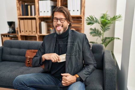 Foto de Handsome middle age man at consultation office keeping money sticking tongue out happy with funny expression. - Imagen libre de derechos