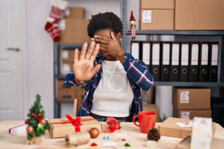 Téléchargez les photos : Femme afro-américaine travaillant dans une petite entreprise faisant décoration de Noël couvrant les yeux avec les mains et faisant un geste d'arrêt avec expression triste et peur. concept embarrassé et négatif. - en image libre de droit