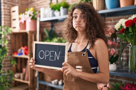 Photo for Hispanic woman with curly hair working at florist holding open sign puffing cheeks with funny face. mouth inflated with air, catching air. - Royalty Free Image