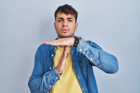 Foto de Joven hombre hispano de pie sobre fondo azul haciendo un gesto de tiempo fuera con las manos, rostro frustrado y serio - Imagen libre de derechos
