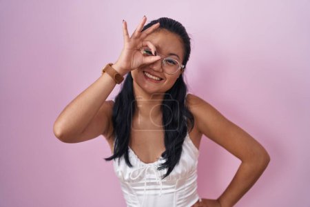 Foto de Asiática joven mujer de pie sobre rosa fondo sonriendo feliz haciendo ok signo con la mano en el ojo mirando a través de los dedos - Imagen libre de derechos