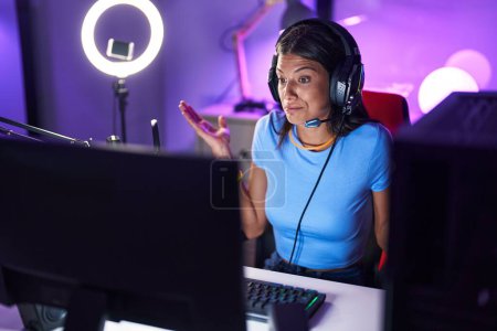 Foto de Brunette young woman playing video games clueless and confused expression with arms and hands raised. doubt concept. - Imagen libre de derechos