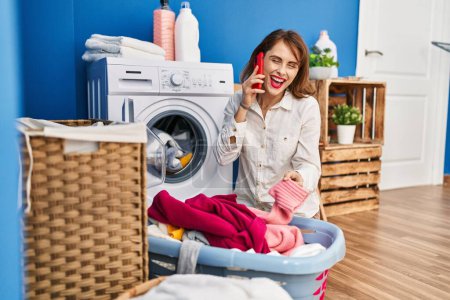 Foto de Mujer joven caucásica hablando en el teléfono inteligente a la espera de la lavadora en la sala de lavandería - Imagen libre de derechos