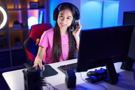 Foto de Joven mujer china streamer sonriendo confiado sentado en la cama en la sala de juegos - Imagen libre de derechos