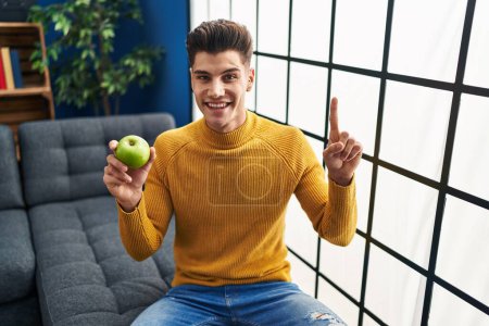 Foto de Joven hispano sosteniendo manzana verde sonriendo con una idea o pregunta apuntando con el dedo con cara feliz, número uno - Imagen libre de derechos