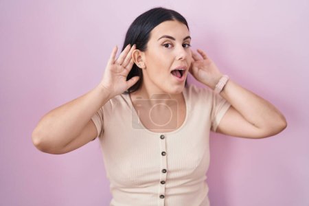 Foto de Joven mujer hispana de pie sobre fondo rosa tratando de escuchar ambas manos en el gesto de la oreja, curiosa de chismes. problema auditivo, sordo - Imagen libre de derechos