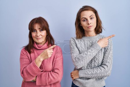 Photo for Mother and daughter standing over blue background pointing with hand finger to the side showing advertisement, serious and calm face - Royalty Free Image