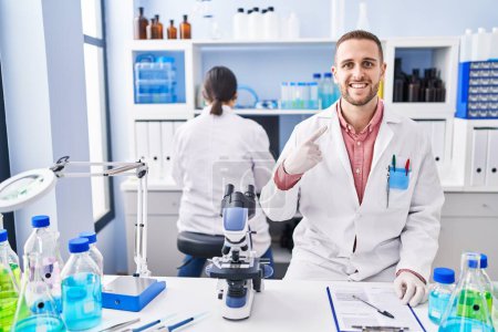 Téléchargez les photos : Jeune homme travaillant au laboratoire scientifique souriant heureux pointant avec la main et le doigt - en image libre de droit