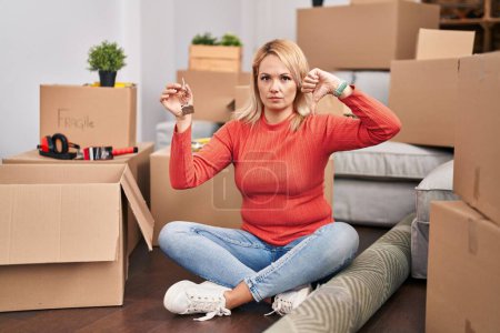 Photo for Blonde woman holding keys of new home sitting on the floor with angry face, negative sign showing dislike with thumbs down, rejection concept - Royalty Free Image