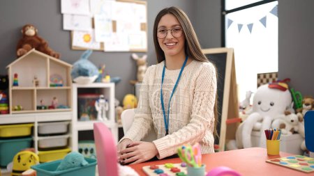 Photo for Young beautiful hispanic woman preschool teacher smiling confident sitting on table at kindergarten - Royalty Free Image