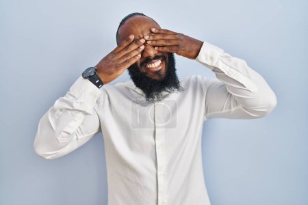 Foto de African american man standing over blue background covering eyes with hands smiling cheerful and funny. blind concept. - Imagen libre de derechos