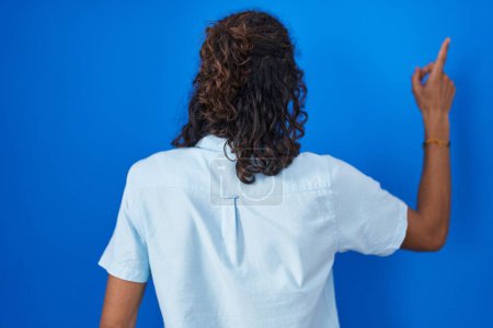 Téléchargez les photos : Jeune homme hispanique debout sur fond bleu posant vers l'arrière pointant vers l'avant avec la main du doigt - en image libre de droit