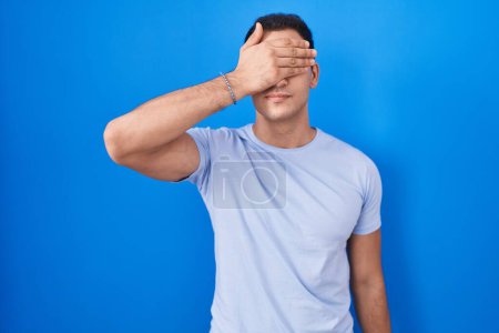 Foto de Young hispanic man standing over blue background covering eyes with hand, looking serious and sad. sightless, hiding and rejection concept - Imagen libre de derechos
