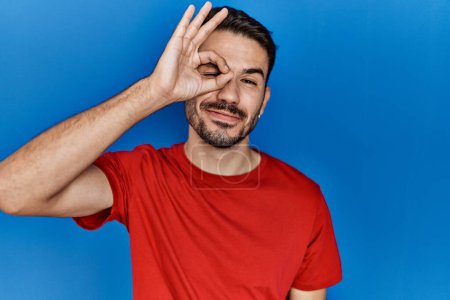 Téléchargez les photos : Young hispanic man with beard wearing red t shirt over blue background doing ok gesture with hand smiling, eye looking through fingers with happy face. - en image libre de droit