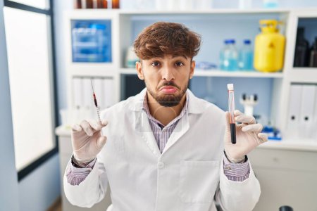Foto de Hombre árabe con barba trabajando en un laboratorio científico sosteniendo muestras de sangre deprimido y preocupado por la angustia, llorando enojado y asustado. expresión triste. - Imagen libre de derechos