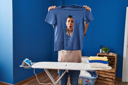 Téléchargez les photos : Jeune homme latino tenant t-shirt avec trou brûlé avec expression surprise à la buanderie - en image libre de droit