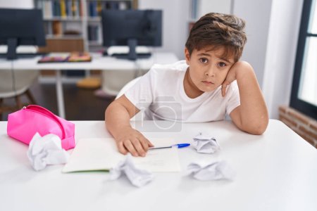 Photo for Adorable hispanic boy student sitting on table with worried expression at classroom - Royalty Free Image