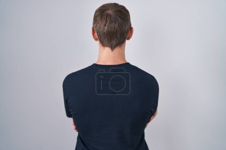 Téléchargez les photos : Blanc blond homme portant Bitcoin t-shirt debout vers l'arrière regardant loin avec les bras croisés - en image libre de droit