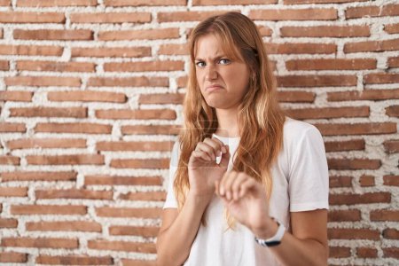 Foto de Young caucasian woman standing over bricks wall disgusted expression, displeased and fearful doing disgust face because aversion reaction. - Imagen libre de derechos