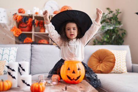 Foto de Adorable chica hispana vestida con disfraz de halloween sentada en un sofá con expresión ganadora en casa - Imagen libre de derechos