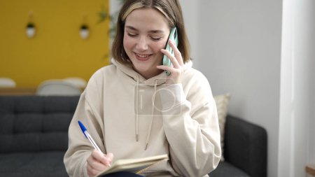 Foto de Mujer rubia joven escribiendo en el cuaderno hablando en el teléfono inteligente en casa - Imagen libre de derechos