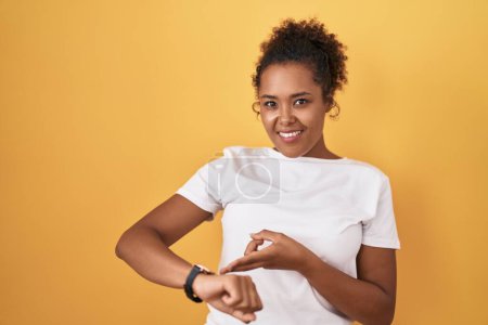 Téléchargez les photos : Young hispanic woman with curly hair standing over yellow background in hurry pointing to watch time, impatience, upset and angry for deadline delay - en image libre de droit