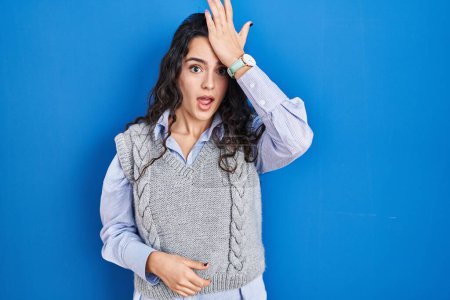 Photo for Young brunette woman standing over blue background surprised with hand on head for mistake, remember error. forgot, bad memory concept. - Royalty Free Image