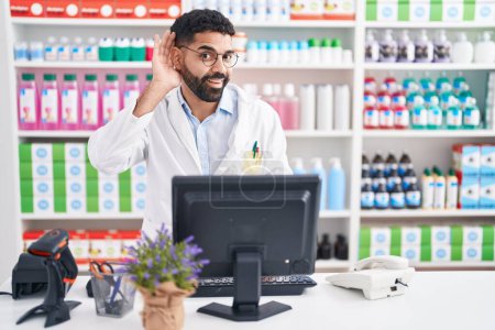 Foto de Hispanic man with beard working at pharmacy drugstore smiling with hand over ear listening an hearing to rumor or gossip. deafness concept. - Imagen libre de derechos