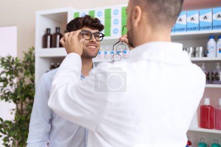 Photo for Two hispanic men pharmacist speaking to client choosing glasses at pharmacy - Royalty Free Image