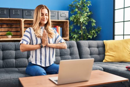 Téléchargez les photos : Jeune femme blonde ayant cours de yoga en ligne assis sur le canapé à la maison - en image libre de droit