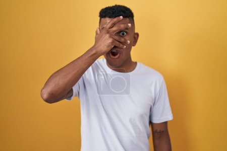 Photo for Young hispanic man standing over yellow background peeking in shock covering face and eyes with hand, looking through fingers with embarrassed expression. - Royalty Free Image