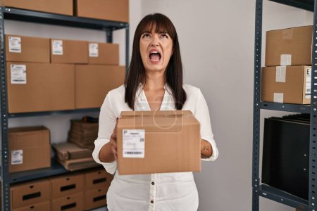 Photo for Young brunette woman working at small business ecommerce holding package angry and mad screaming frustrated and furious, shouting with anger looking up. - Royalty Free Image
