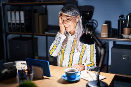 Foto de Mujer de mediana edad con el pelo gris trabajando en la oficina por la noche cubriendo un ojo con la mano, sonrisa confiada en la cara y emoción sorpresa. - Imagen libre de derechos