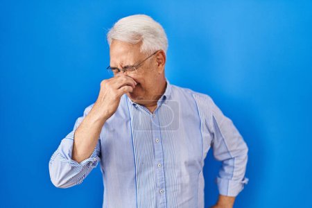 Téléchargez les photos : Homme âgé hispanique portant des lunettes sentant quelque chose de puant et dégoûtant, odeur intolérable, retenant son souffle avec les doigts sur le nez. mauvaise odeur - en image libre de droit