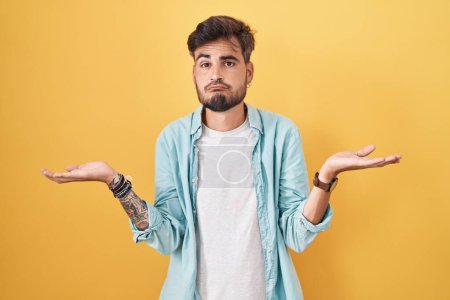 Foto de Young hispanic man with tattoos standing over yellow background clueless and confused expression with arms and hands raised. doubt concept. - Imagen libre de derechos