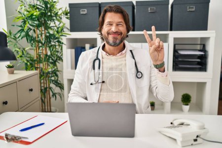 Foto de Handsome middle age doctor man working at the clinic showing and pointing up with fingers number two while smiling confident and happy. - Imagen libre de derechos