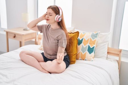 Foto de Young redhead woman doing yoga exercise sitting on bed at bedroom - Imagen libre de derechos