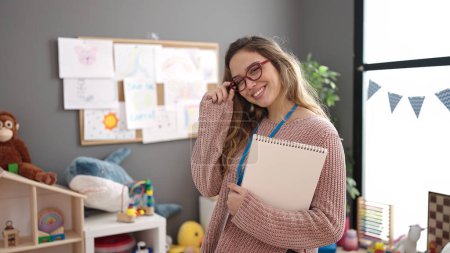 Téléchargez les photos : Young beautiful hispanic woman preschool teacher smiling confident holding notebook at kindergarten - en image libre de droit