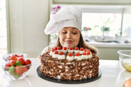 Foto de Joven hermosa mujer hispana sonriendo confiada oliendo pastel en la cocina - Imagen libre de derechos