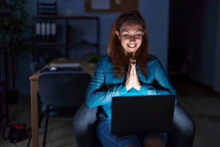 Téléchargez les photos : Brunette woman working at the office at night praying with hands together asking for forgiveness smiling confident. - en image libre de droit