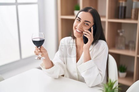 Photo for Young hispanic woman talking on the smartphone and drinking wine at home - Royalty Free Image