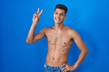Téléchargez les photos : Jeune homme hispanique debout torse nu sur fond bleu souriant en regardant la caméra montrant des doigts faisant signe de victoire. numéro deux. - en image libre de droit