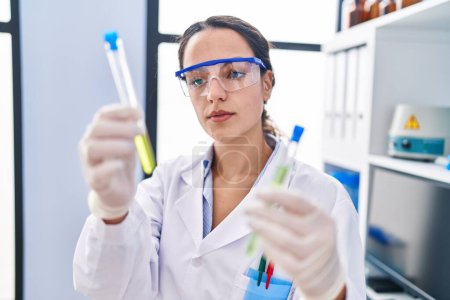 Foto de Mujer hispana joven vistiendo uniforme científico sosteniendo tubos de ensayo en laboratorio - Imagen libre de derechos