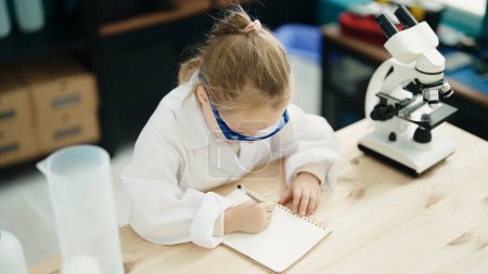 Photo for Adorable blonde girl student using microscope writing on notebook at laboratory classroom - Royalty Free Image