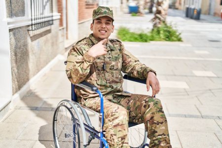 Téléchargez les photos : Jeune homme arabe portant un uniforme de camouflage de l'armée assis sur fauteuil roulant souriant heureux pointant avec la main et le doigt - en image libre de droit