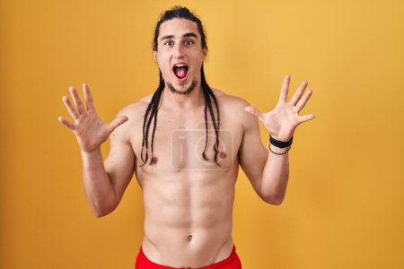 Téléchargez les photos : Homme hispanique avec les cheveux longs debout torse nu sur fond jaune célébrant fou et étonné pour le succès avec les bras levés et les yeux ouverts hurlant excités. concept gagnant - en image libre de droit